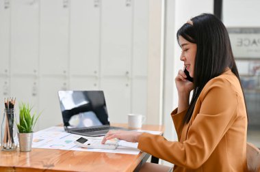 Güzel ve çekici Asyalı genç iş kadını ya da muhasebeci ofis masasında çalışıyor ve iş müşterisiyle telefon görüşmesi yapıyor..