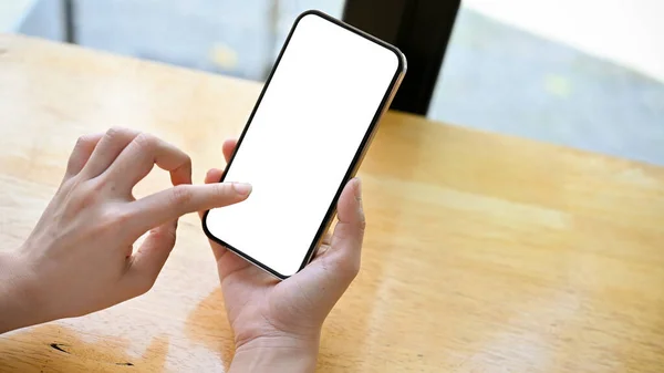 A smartphone white screen mockup is on a woman's hand over the table. top view, Female using touchscreen smartphone.