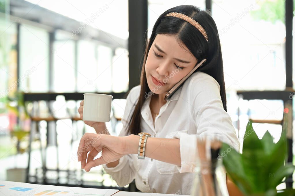 Busy and professional millennial Asian female CEO or businesswoman at her office desk, having a morning coffee, checking time on her wristwatch while talking on the phone.