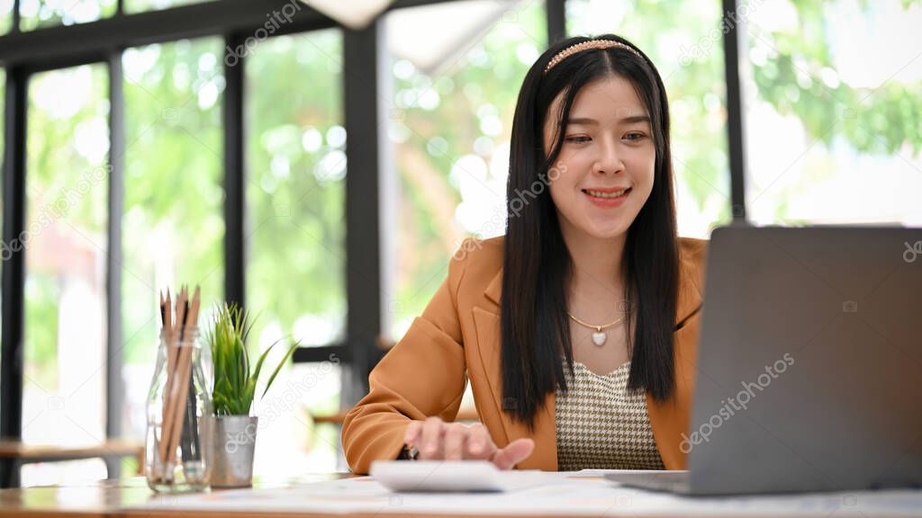 Beautiful Asian female accountant or financial worker sits at her desk, working on her financial task on laptop computer.