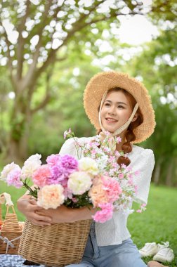 Portre, güzel ve çekici Asyalı kadın hasır şapka takıyor ve elinde çiçek sepeti yeşil bahçede piknik yapıyor..
