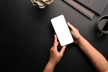 Overhead view, A female hands holding a smartphone white screen mockup over her modern black workspace.