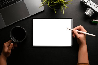 Modern siyah ofis masasında dijital tablet kullanan, kahve fincanı ve stilus kalem tutan bir kadın el. Tablet beyaz ekran modeli. en üstteki görünüm