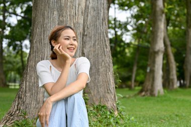 Çekici ve mutlu Asyalı genç bayan bir parkın güzel yeşilliklerinde bir ağacın altında rahatlıyor..