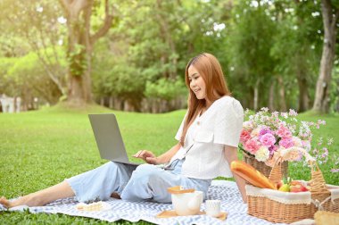 Çekici genç Asyalı bayan ya da üniversite öğrencisi yeşillik parkında piknik yapıyor, piknik örtüsünün üzerinde oturuyor ve okul ödevini yapmak için taşınabilir dizüstü bilgisayar kullanıyor..