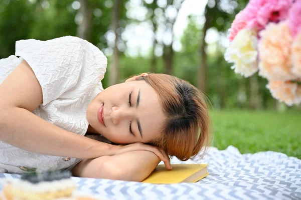 Relaxed and calm young Asian female laying down on a picnic blanket, taking a nap in sunny picnic day in the green park.