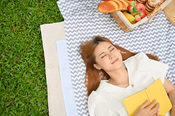 Top view, Beautiful and attractive young Asian female laying down on her picnic blanket with her favourite book. Picnic concept