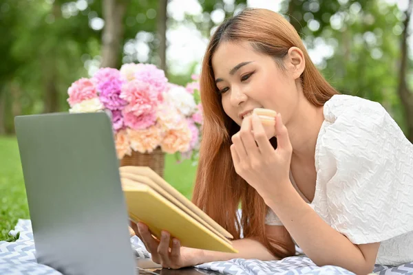 Attractive young Asian female laying down on picnic blanket, reading a book, eating some sweet and having a nice picnic day in the garden.