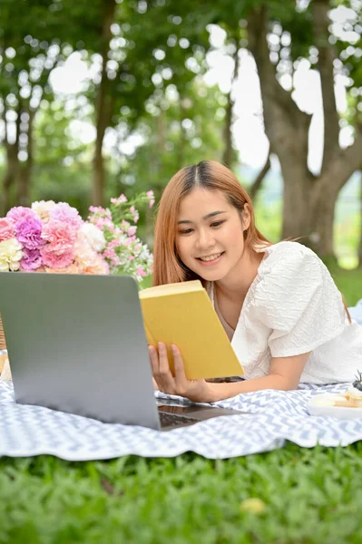 Beautiful young Asian female laying on picnic blanket, reading a book and having a nice picnic day in the garden.