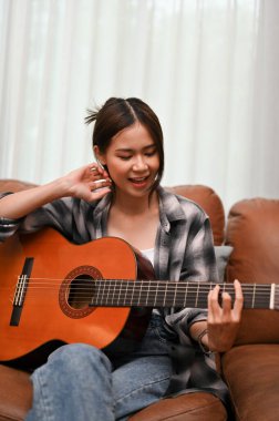 Portrait, An attractive and charming young Asian female is on the sofa in her living room, singing and playing guitar. Creative hobby concept.