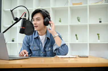 Talented and smart young Asian male radio host or VJ wearing headphones and speaking on his professional microphone, running his podcast in studio.