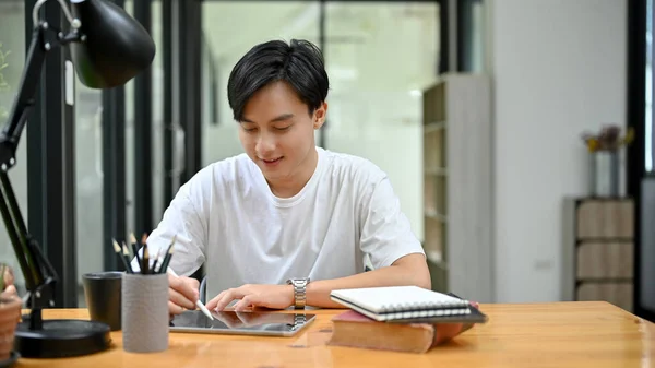 Handsome young Asian male university student concentrating doing his homework on tablet touchpad in the library co-working space.