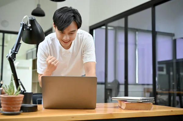 Handsome young Asian man looking at laptop screen, feeling happy after getting a wonderful email, job promotion, passing the exam, winning the lottery, or project success.