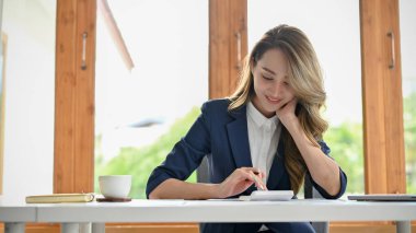 Attractive young Asian female accountant or businesswoman sits at her office desk, working on her financial task and using calculator.