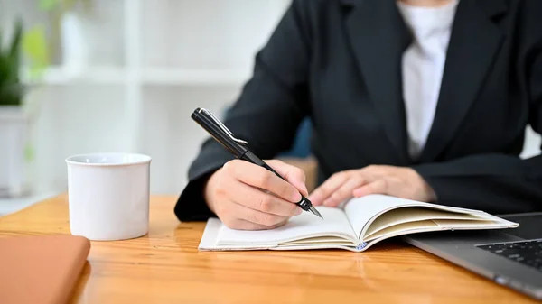 A businesswoman or female lawyer in formal black suit writing something on her notebook at the office desk. cropped image