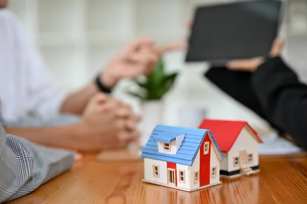 A house model is on the meeting table over a blurred background with a real estate agent and client having a meeting. cropped image