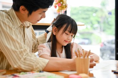 Proud Asian dad patting on his daughter's head while teaching her homework at home. Lovely family concept