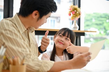 Happy and adorable young Asian girl giving thumbs up to her teacher after finished homework or class.