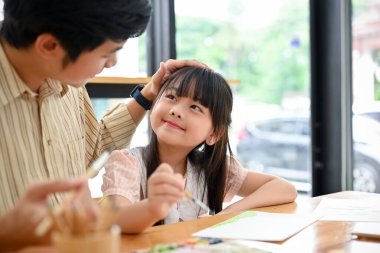 Happy proud Asian dad and daughter having fun together, enjoying activity, drawing and painting watercolor together at home.