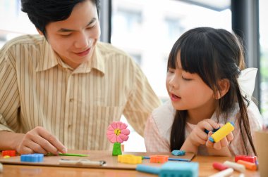 Smiling and proud Asian dad and lovely daughter playing play dough at home, moulding colorful clay or plasticine together. Happy family time concept