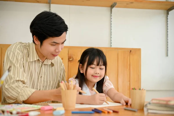 A handsome Asian dad and a cute daughter enjoy drawing and painting a rainbow on paper together at home. Dad and daughter activity concept.