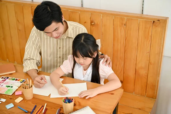 Professional and kind Asian male art teacher or tutor teaching a cute young girl student to draw and paint with watercolor. above view