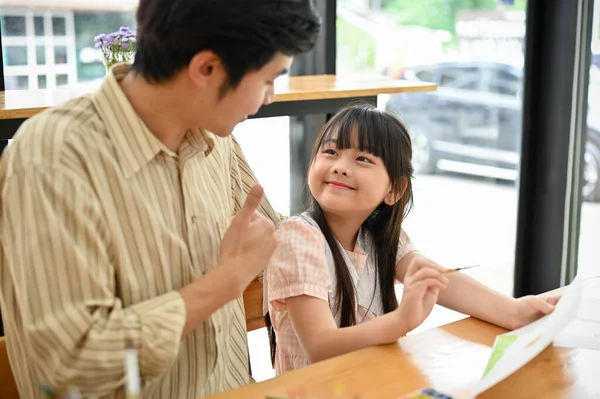 A proud Asian dad gives a thumbs up to his cute daughter while teaching art to her. Happy and smiling daughter is enjoying a painting workshop with her father.