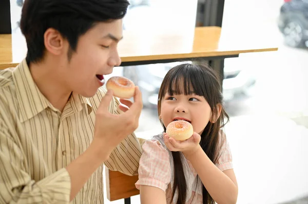 Kind and happy Asian dad enjoys eating doughnuts with her cute young daughter in the coffee shop. Happy family time.