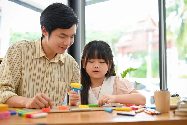 A proud and kind Asian male art teacher is teaching a young girl to sculpt and mould plasticine into different shapes.