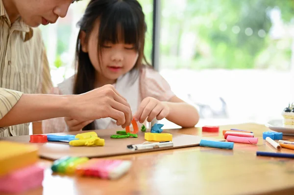 Happy Asian dad and daughter enjoys moulding colorful clay or play dough together at home. selective focus on objects. cropped image