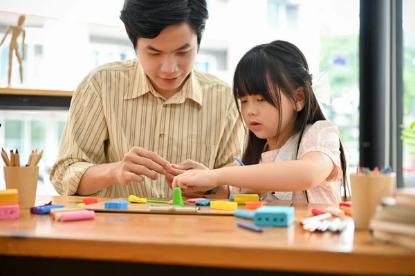 Smiling Asian male art teacher and a young girl moulding colorful clay or plasticine together at school playroom. Creative kids' activity concept.