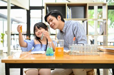 Cute Asian girl enjoy learning science with her male teacher, holding a test tubes, doing an easy experiment for kids.