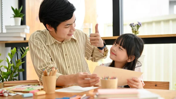 Handsome and kind Asian male art teacher giving a thumbs up to his adorable young student. Asian dad and daughter enjoys painting together.