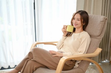 Güzel ve rahat genç Asyalı kadın oturma odasında rahat koltuğunda otururken kahvesini yudumluyor..
