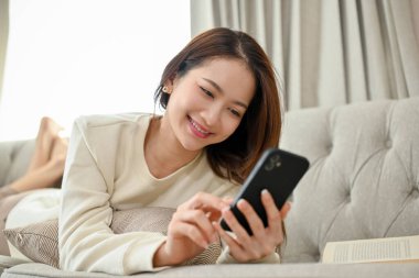Çekici genç Asyalı kadın rahat koltuğunda uzanıyor, sosyal medyada kayıyor ve akıllı telefonuyla arkadaşlarıyla sohbet ediyor..