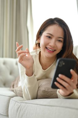 Portre, çekici ve mutlu genç Asyalı kadın rahat koltuğunda uzanıyor, sosyal medyada kayıyor ve akıllı telefonuyla arkadaşlarıyla sohbet ediyor..