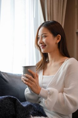 Çekici ve muhteşem Asyalı kadın oturma odasında rahat bir koltukta oturur, kahvesini yudumlar ve pencereden dışarı bakar, mutluluğu hayal eder..