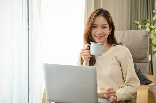 Beautiful millennial Asian female relaxes sits in her living room, sipping morning coffee and using laptop computer.