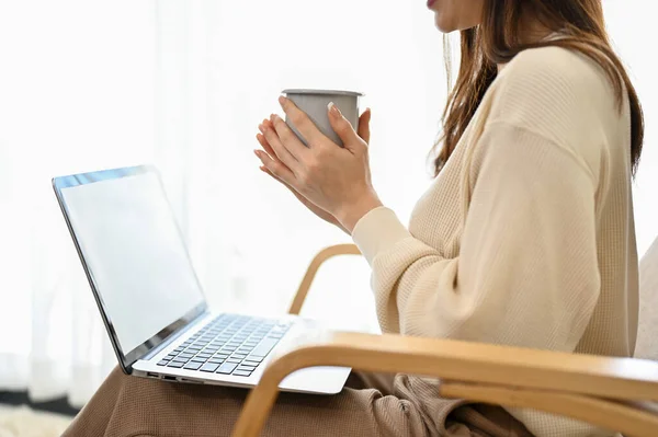 Relaxed and happy young Asian female sits on armchair in the living room, holding a hot coffee mug and using laptop computer. close-up and cropped image