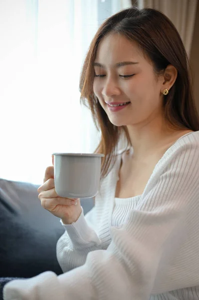 Portrait, Attractive and charming young Asian female in the living room holding a cup of coffee, sipping a morning coffee to start her weekend.