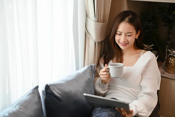 美丽迷人的年轻亚洲女性在舒适的客厅里躺在沙发上 一边喝着热咖啡 一边用数码平板电脑 — 图库照片