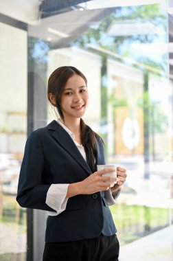 Çekici Asyalı genç bir iş kadını ya da takım elbiseli bir kadın patronun portresi cam pencerenin yanında duruyor ve elinde bir fincan kahve tutuyor..