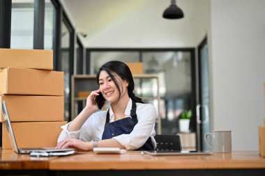 Çekici Asyalı genç bayan çevrimiçi dükkan sahibi, ya da ofiste e-ticaret girişimcisi, müşterisiyle telefonda konuşuyor..