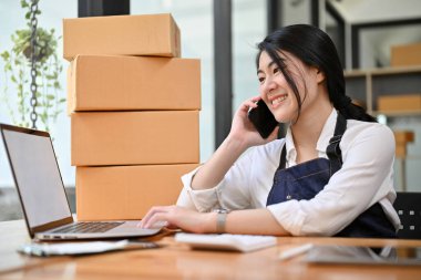 Genç ve güzel Asyalı kadın e-ticaret girişimcisi. Tedarikçisiyle telefonda konuşurken dizüstü bilgisayarındaki ürün hisselerine bakıyor..