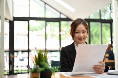 Resmi takım elbiseli güzel Asyalı bayan ofis çalışanı ofis çalışma alanındaki kağıt üzerinde iş bilgilerini okumaya odaklanmış..