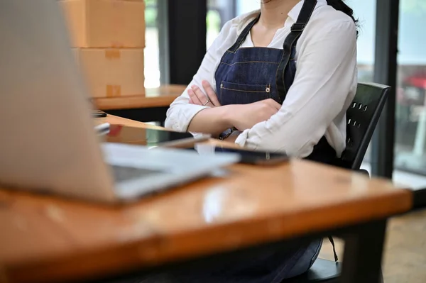 A professional young Asian female entrepreneur or small online business owner sits at her office desk with arms crossed. cropped image