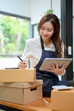 Muhteşem genç Asyalı kadın internet işi girişimi Sevkiyat paketini kontrol ediyor ve dijital tableti tutuyor.