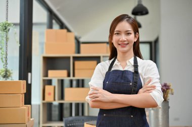 Portre, başarılı ve kendinden emin genç Asyalı kadın e-ticaret işyeri sahibi ya da girişimci ofiste kollarını kavuşturmuş duruyor.