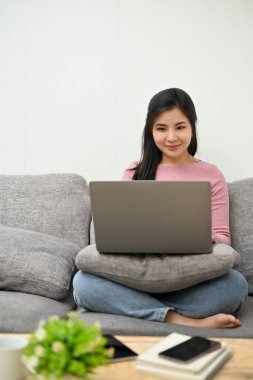 Asyalı çekici bir kadın oturma odasındaki kanepede dizüstü bilgisayarla online işlerini hallediyor..