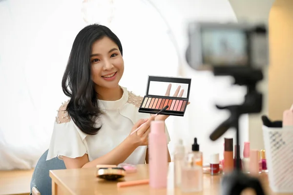 Beautiful young Asian female beauty blogger or makeup artist recording makeup tutorial video or live streaming on her online platform in her living room.